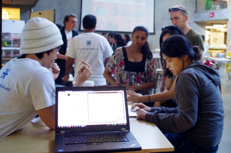 A few people sits around a desk, discussing ideas. A laptop is propped open at the end of the table, its white screen shows some lines of code. Some people in the background can be seen moving around.