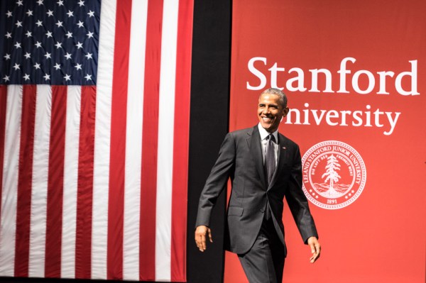 Former President Barack Obama walking in front of an American flag and a wall reading "Stanford University"