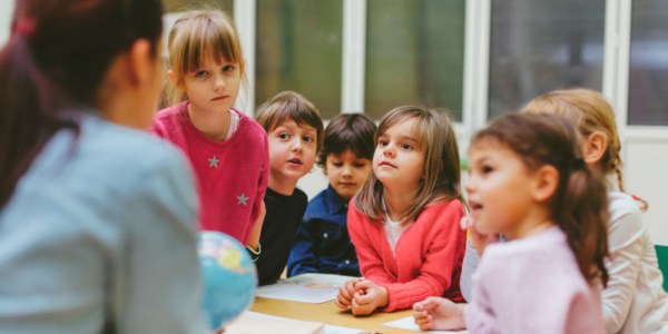 Preschool teacher teaching her children about geography. Using globe and asking the questions. Children  answering the questions. Children sitting by the table. Selective focus to girl in the middle. Models in this shot are part of real kindergarten group and their teacher.