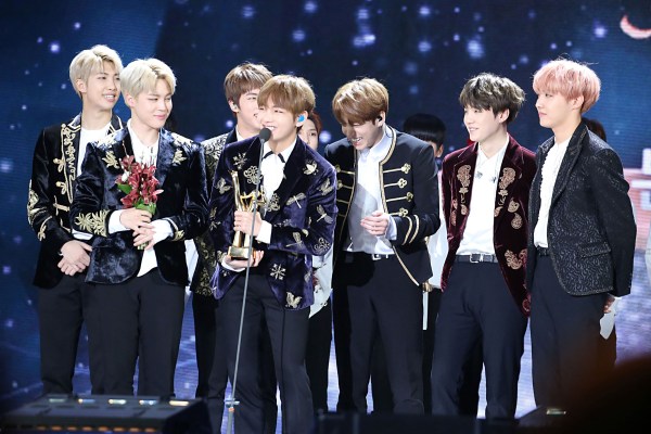 BTS stands in white shirts and black jackets on a stage holding a golden disk award.