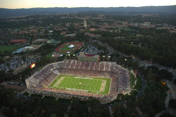 Stanford Stadium seen from the sky at dusk. The stadium is lit, with players on the field and spectators in the stands