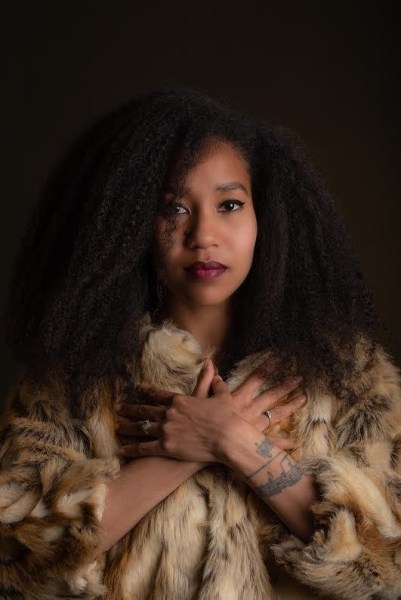 Minhee Jones stands against a dark, solid background in a fur coat. There is a musical staff tattooed on her left arm.
