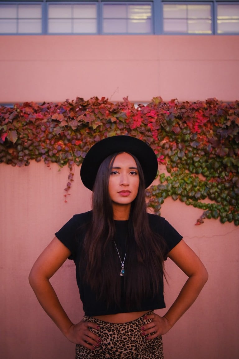 Raye Zaragoza stands with her hands on her hips against a light-colored wall with leaves on it. She wears a broad-rimmed black hat, short black tee and a cheetah print skirt.