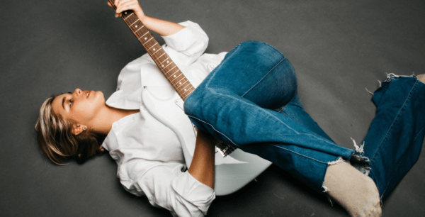 Alex McArtor lays on a solid gray floor, holding a white electric guitar. She wears a white button up and blue jeans.