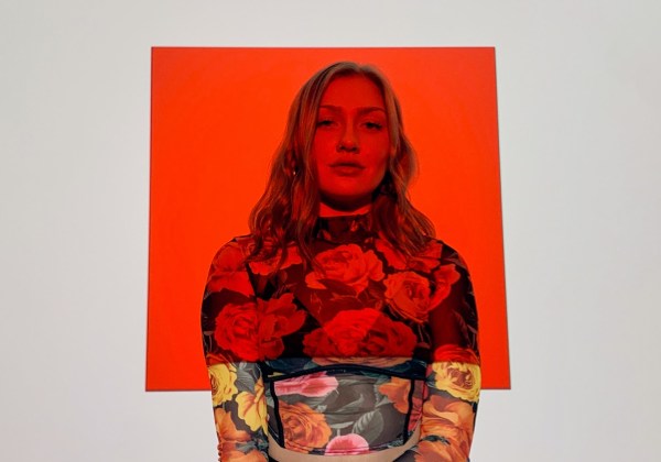 Olivia Lunny stands against an off-white background with a red square projected over her face and upper torso. She wears a sheer floral long-sleeve.