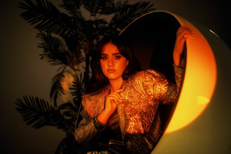 Tia Gostelow sits in a spherical chair, bathed in orange mood lighting. She wears a golden, sequined jacket that catches the light.