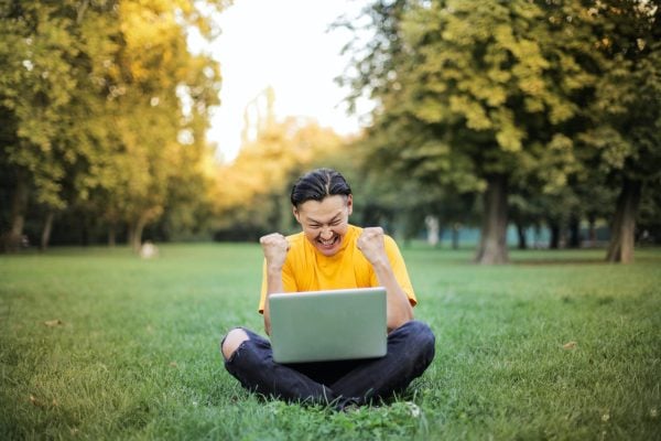 An excited person sitting on grass with a laptop. (Photo by Andrea Piacquadio from Pexels)
