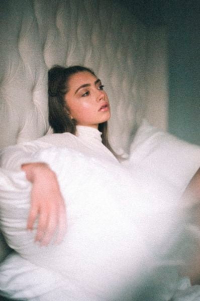 Baker Grace leans against a white headboard, resting her arms on two white pillows. She also wears a white shirt.