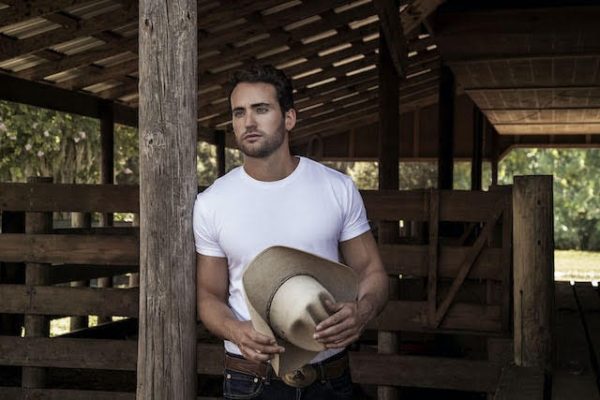 A man in a white T-shirt and jeans stands in a stable holding a cowboy hat.