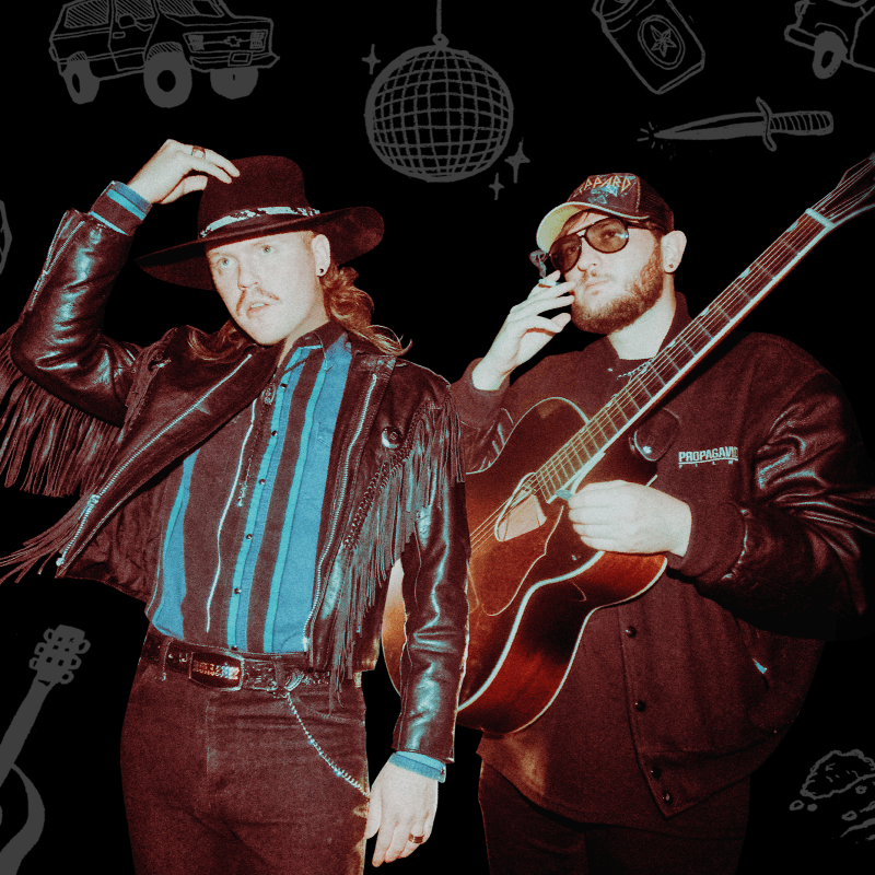 Adam Walker and Vince Winik stand in front of a black background decorated with gray drawings. One is in a fashionable cowboy outfit, while the other holds a guitar.