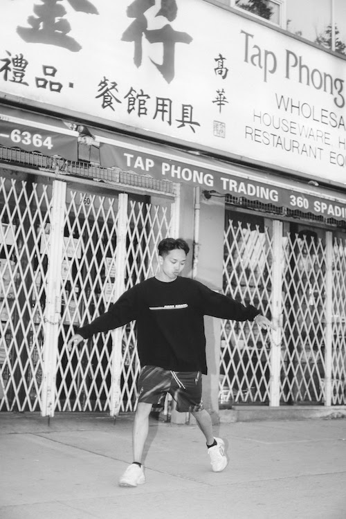 A black and white photo of Nilo Blues in front of Tap Phong Trading. He is wearing a dark sweater and shorts.
