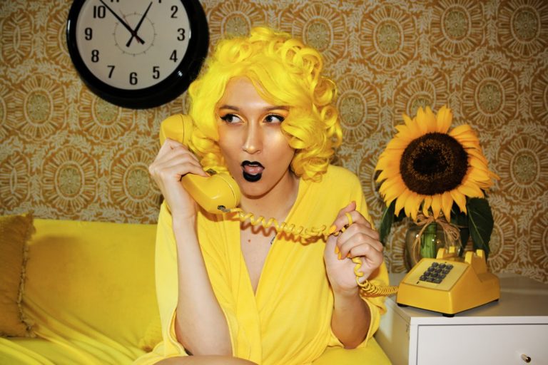 A yellow-tinted woman in a bright yellow dress and matching wig holds a yellow cord phone.