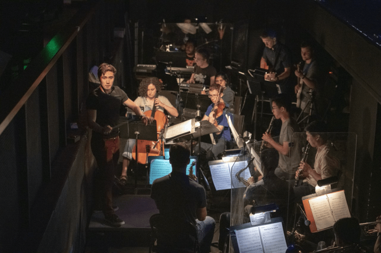 Joshua Chang is conducting a pit orchestra in a dimly lit space