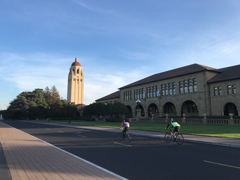 Pictured: two people bike through Stanford campus on a sunny, nearly cloudless day.