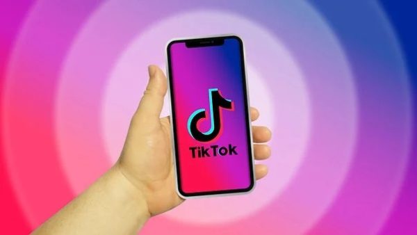Hand holding phone with the Tik Tok app logo displayed on the screen with a pink to purple gradient.