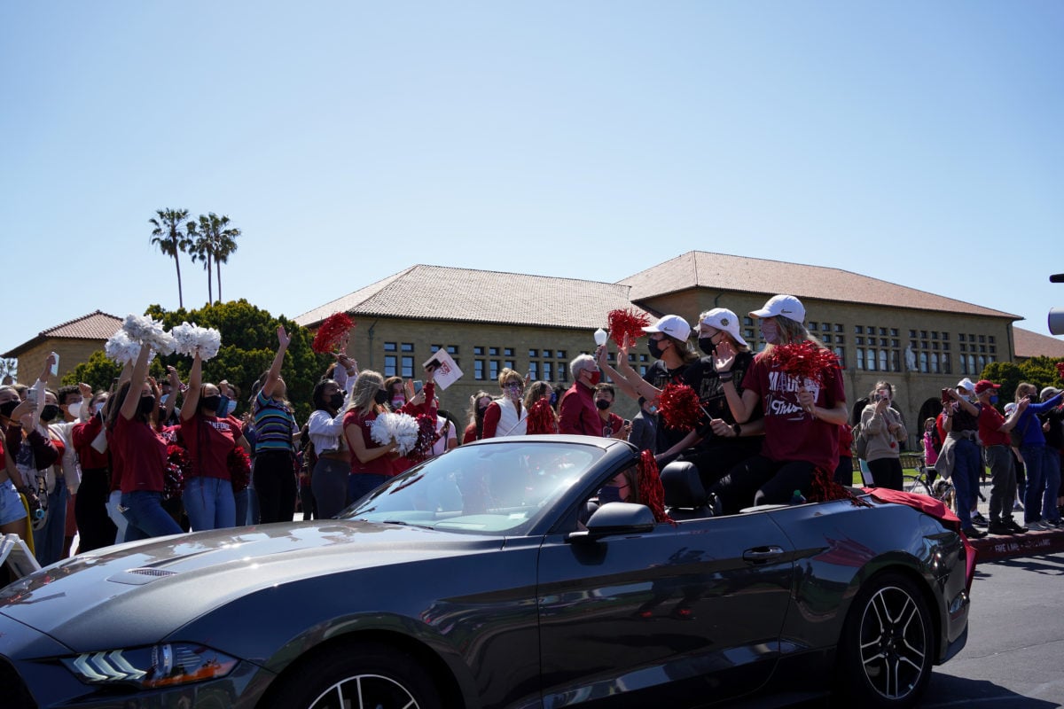 Three women's basketball players sit in a convertible in the Oval as dozens of students, some carrying pom poms, cheer them on.