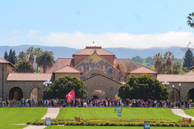 A wide shot of the Oval and Main Quad from Palm Drive. Along a street in the distance about 100 peolpe are gathered and one is waving a giant red flag with Stanford's block 's.'