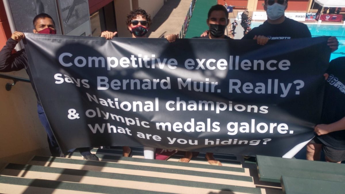 Members of the wrestling team standing in Avery Aquatics Center with white text on a black banner that reads: "Competitive excellence says Bernard Muir. Really? National champions & olympic medals galore. What are you hiding?
