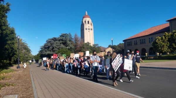 Students march down Palm Drive with Hoover Tower in the background. All are wearing masks and some are wearing signs condemning the Israeli occupation of Palestine.