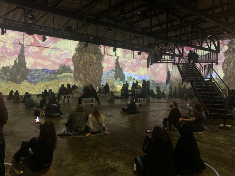 A room full of socially-distanced people looking at a van gogh projection that covers all the room's walls