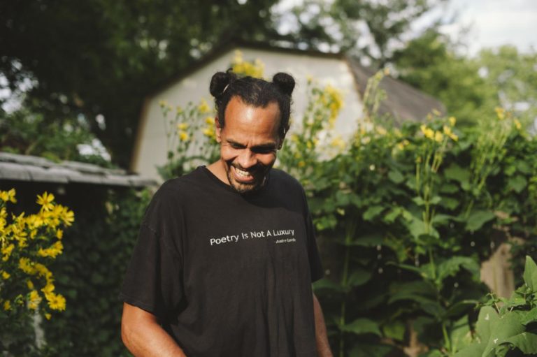 headshot of Ross Gay smiling and looking down, with a shirt that says "Poetry is not a luxury. -Audre Lorde"