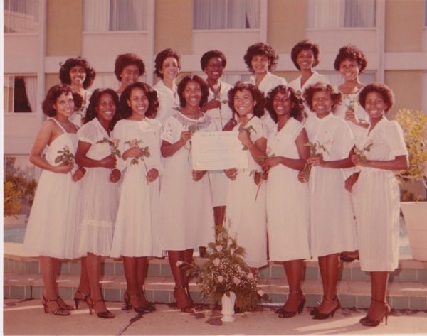 Fifteen women wearing white dresses and brown heels standing in two rows in front of a beige building. Each woman is holding a rose, and the two women in the middle of the front row are holding a certificate.