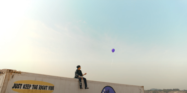 A man wearing all black, sitting on a trailer with "JUST KEEP THE RIGHT VIBE," releasing a purple balloon into the sky.