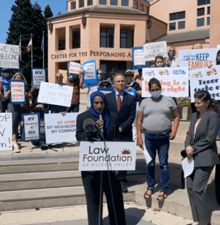 A woman in a black suit and blue hijab speaks into a microphone from behind a podium labeled "Law Foundation of Silicon Valley" and in front of a crowd of people holding signs.