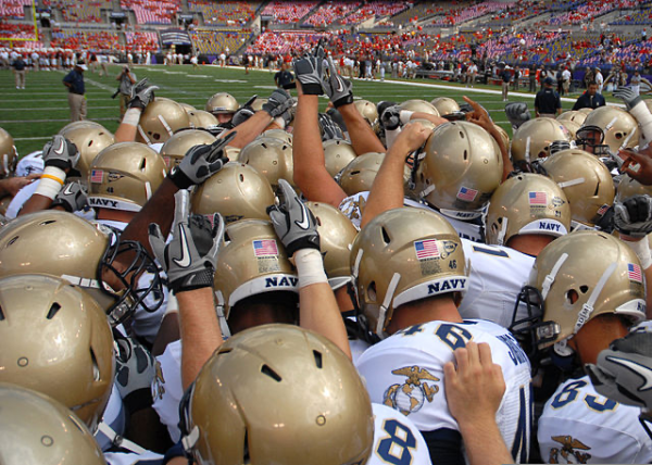 A squad of Navy football players huddles on a field on game day.