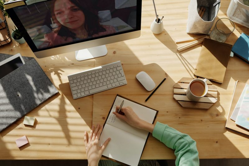 a stock image of someone working at a desk. at top is a computer monitor with a person on video call. at bottom is the person writing in a notebook. elsewhere on the table are a lamp, pencil holder, keyboard and mouse, and coffee.