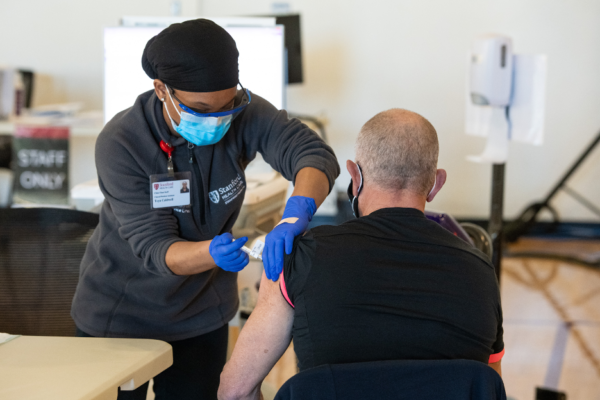 Woman wearing grey Stanford Healthcare sweatshirt and a mask injects COVID-19 vaccine into left arm of man in a black shirt, facing away from the camera.
