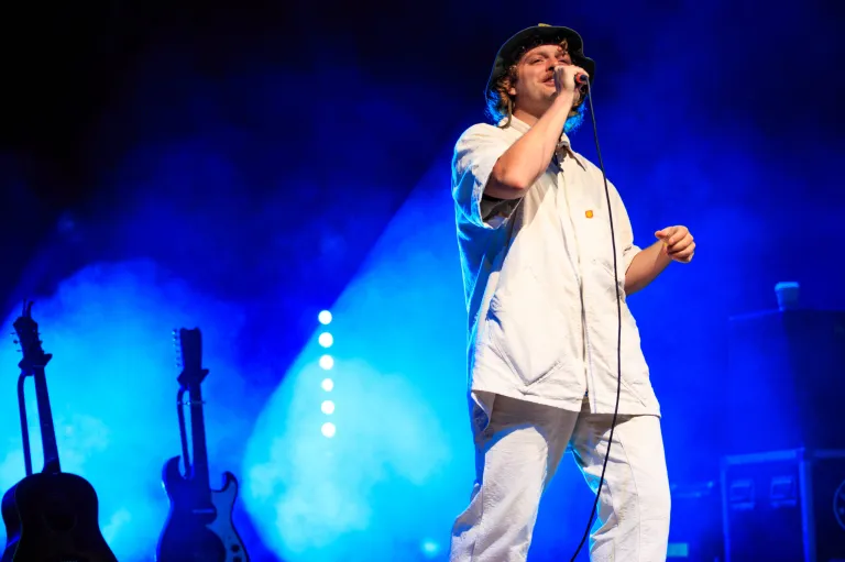 Mac DeMarco on stage singing into a mic with a blue background behind him.