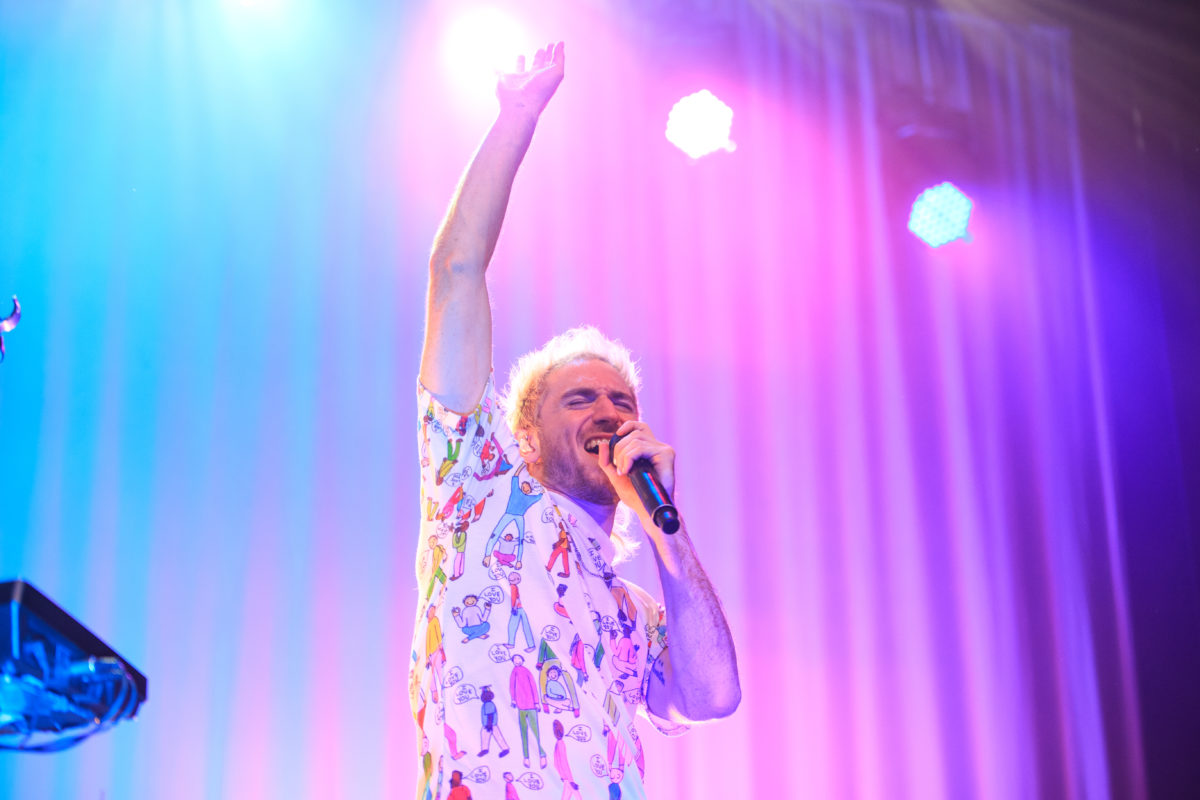 Medium image of Nicholas Petricca singing with his hand stretched upwards on a blue, purple and pink-lit stage.