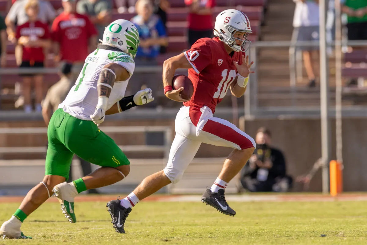 Quarterback Jack West in red and white, holding a football, runs from an Oregon defender on the field. 