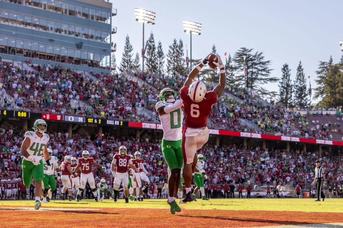 Junior wide receiver Elijah Higgins  jumps up to catch a football in the touchdown zone in front of a full stadium. An Oregon player jumps up next to him to try to catch the ball. Another Oregon player runs towards the two from the left.