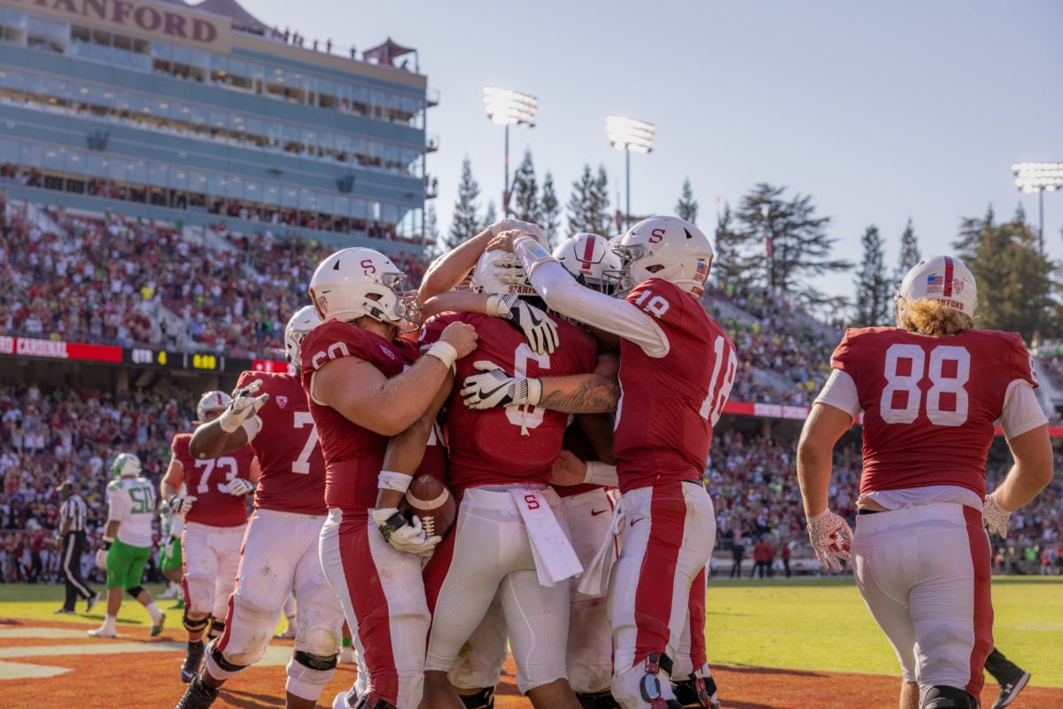Stanford teammates in cardinal red surround wide receiver Elijah Higgins in a group hug at Stanford Stadium. Blurred crowd in the background. 