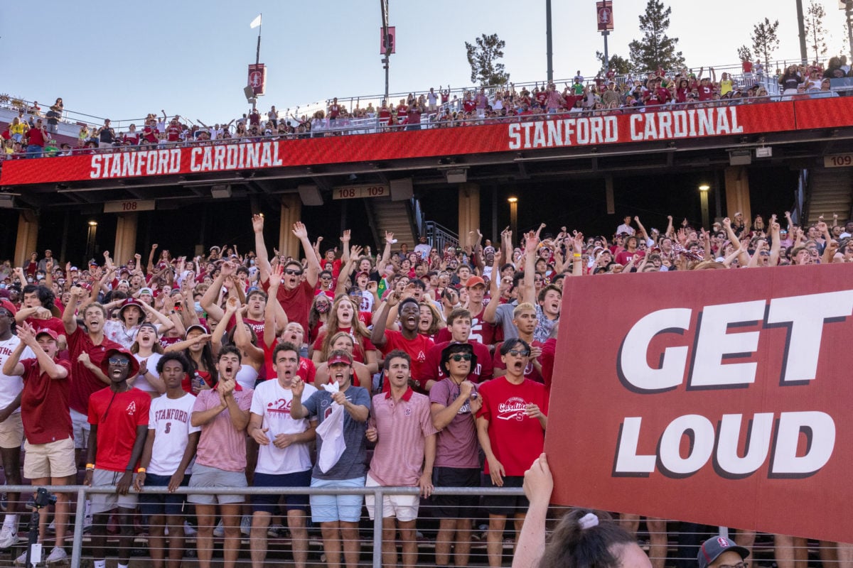 Hundreds of students cheering with their arms outstretched, clad in red, at the Stanford Stadium. A person holds a red sign with white text that says, "GET LOUD."