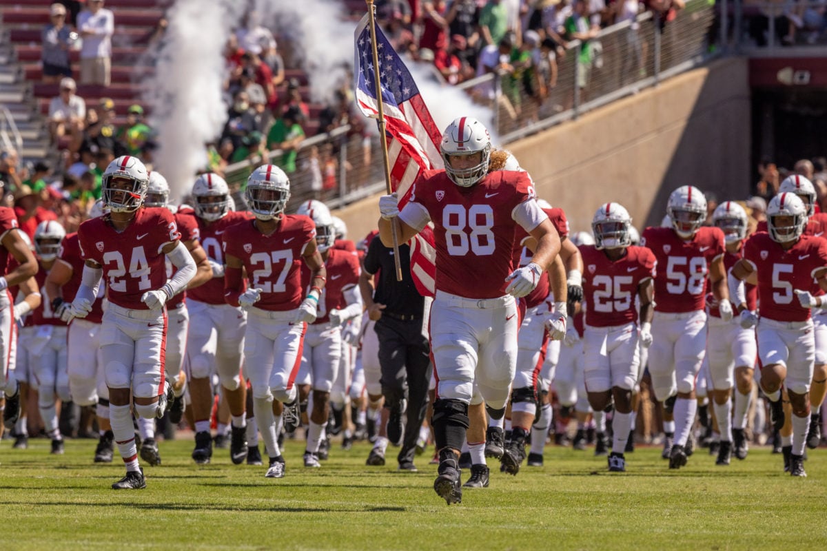 Dozens of football players in red and white running onto the field with Tucker Fisk at the front, carrying the American flag.