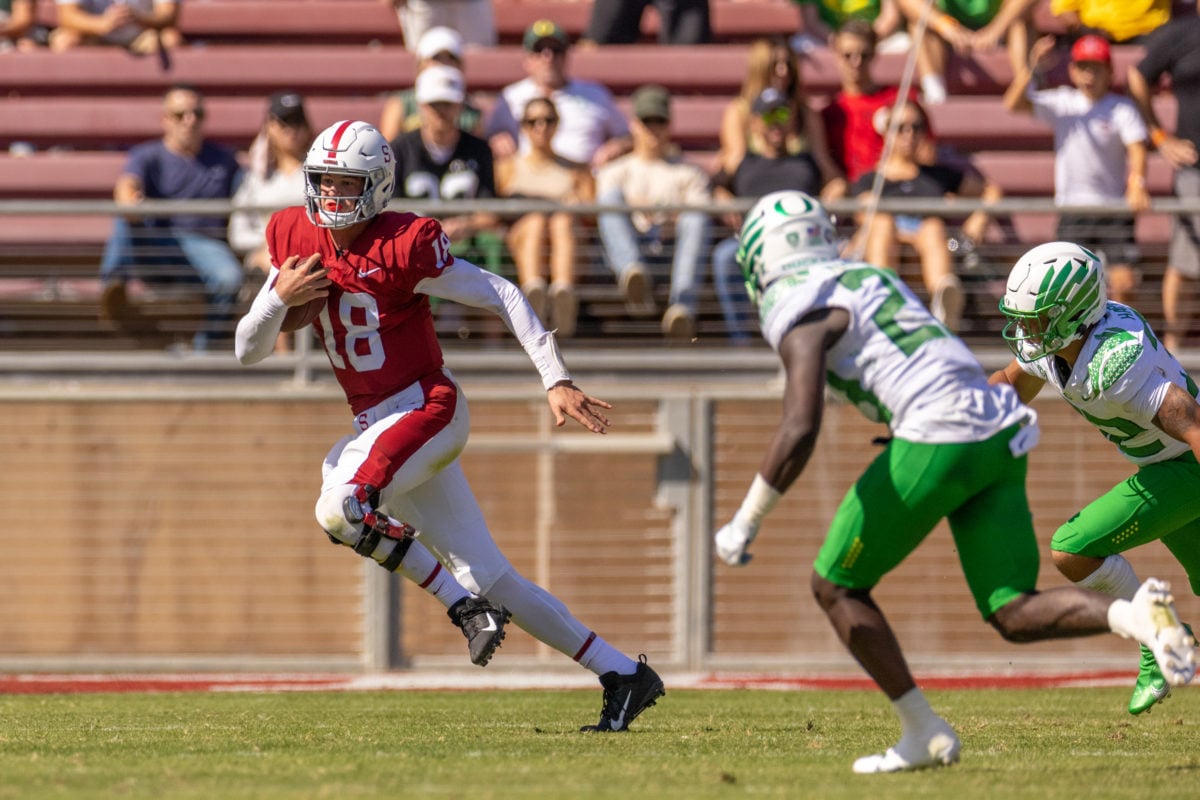 Cardinal quarterback Tanner McKee runs from two Oregon players in green and white. Tanner clutches a football. 