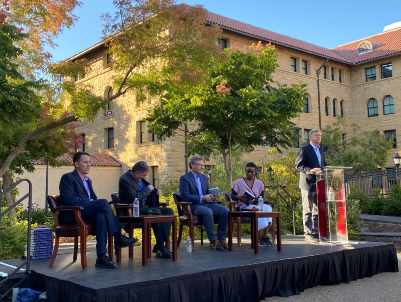 From left to right, Jeremy Weinstein, Mehran Sahami, Rob Reich, Julie Owono and Michael McFaul on an outdoor stage, with McFaul standing at a podium.