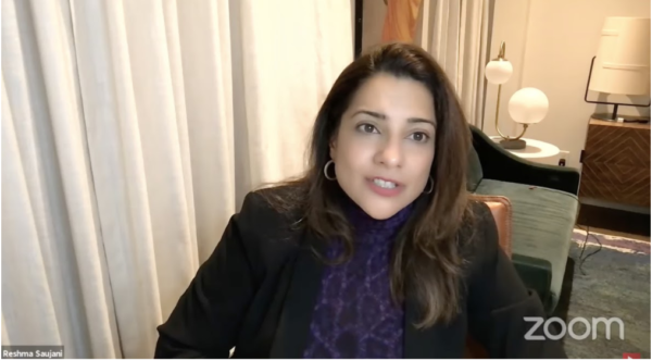 Reshma Saujani over Zoom with white curtains and armchair in the background.