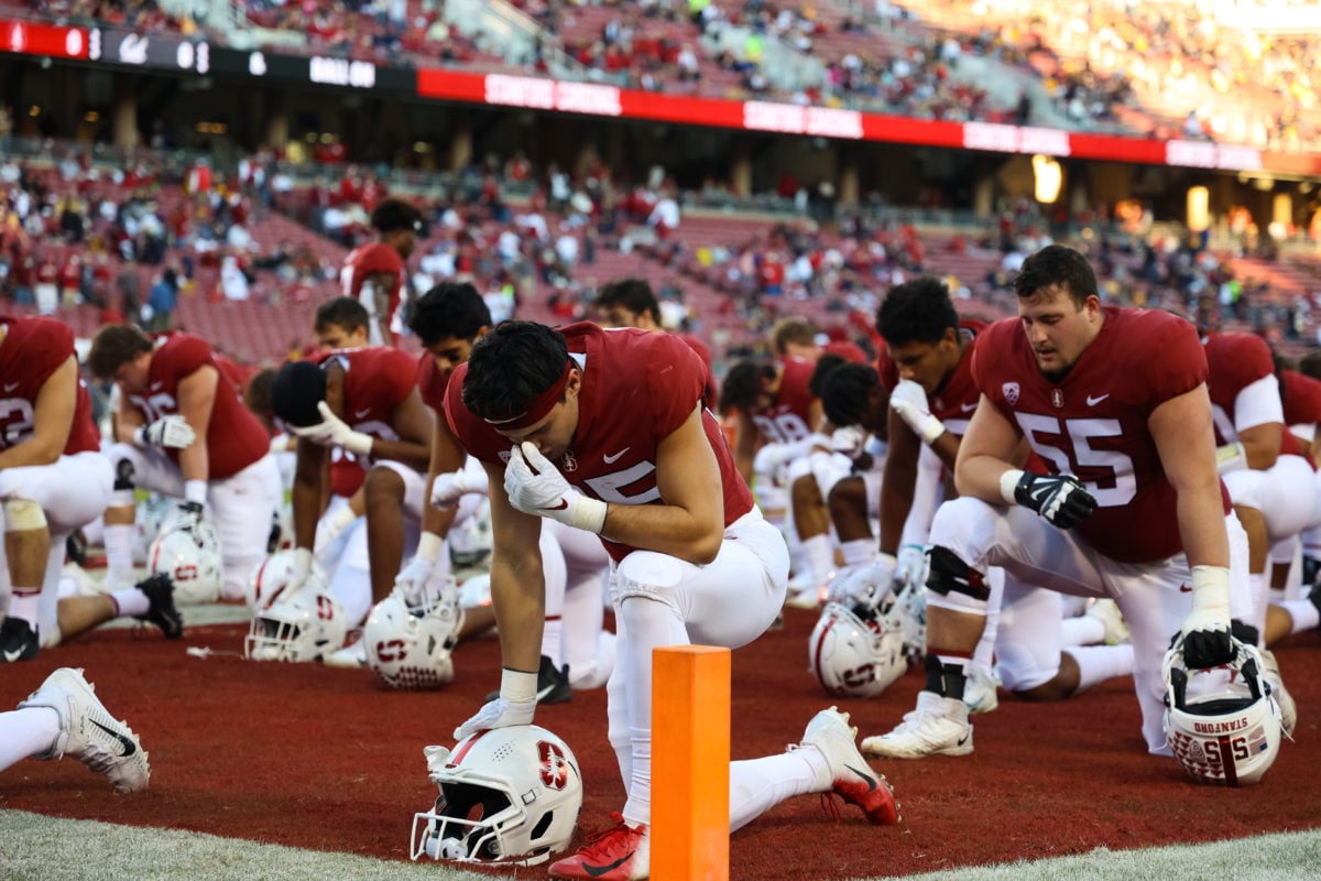 Dozens of Stanford football players kneeling, eyes closed and looking down with their helmets off on the football field