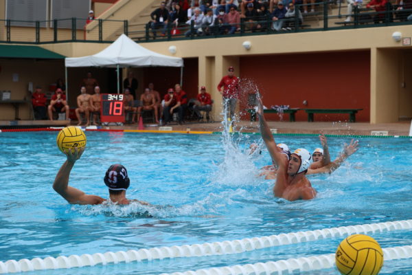A water polo player looks to pass the ball with another player rising out of the water with his hands up to block the pass.