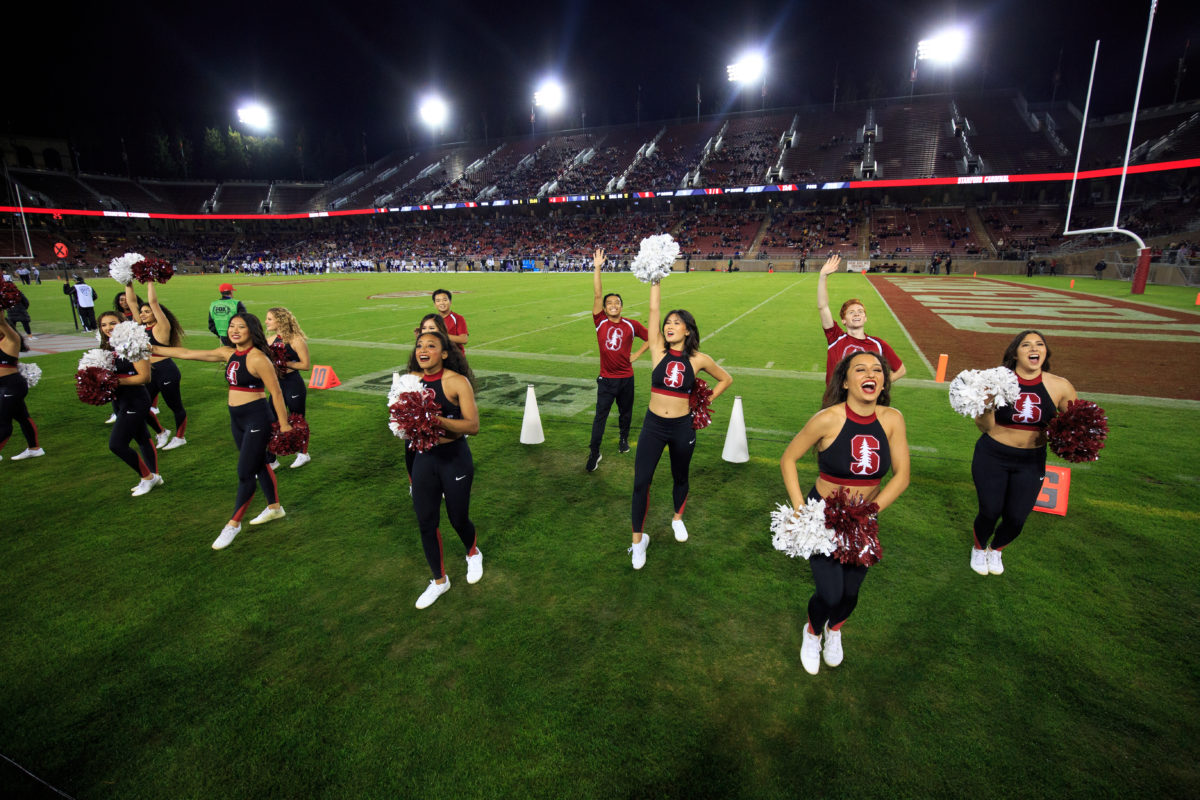 Stanford cheerleading team wave their hands and pom-poms at the crowd on the sidelines of the football field. 