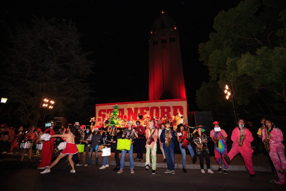 In the night, the Stanford Axe Committee, Football, Band and Cheer dance in front of a banner labeled "Stanford". Hoover Tower is lit red in the background.