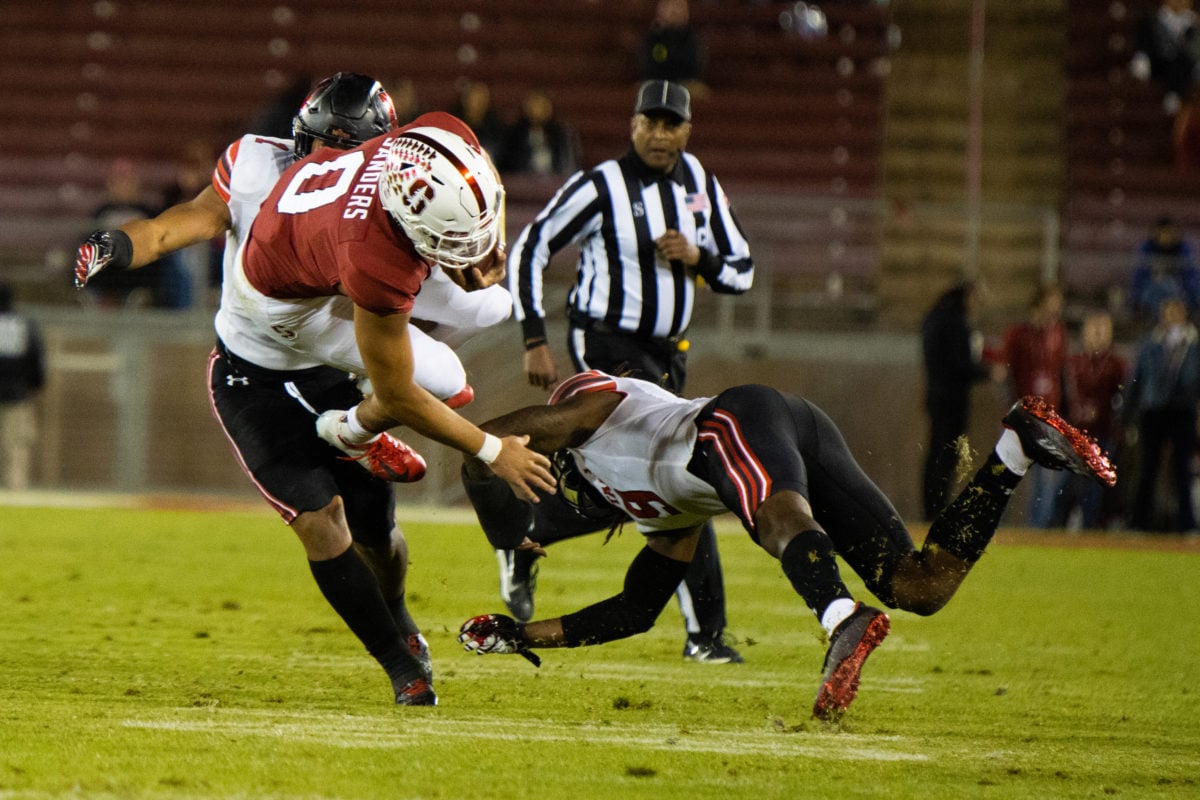 Stanford player in air after Utah players tackle on the field, referee in the background. 