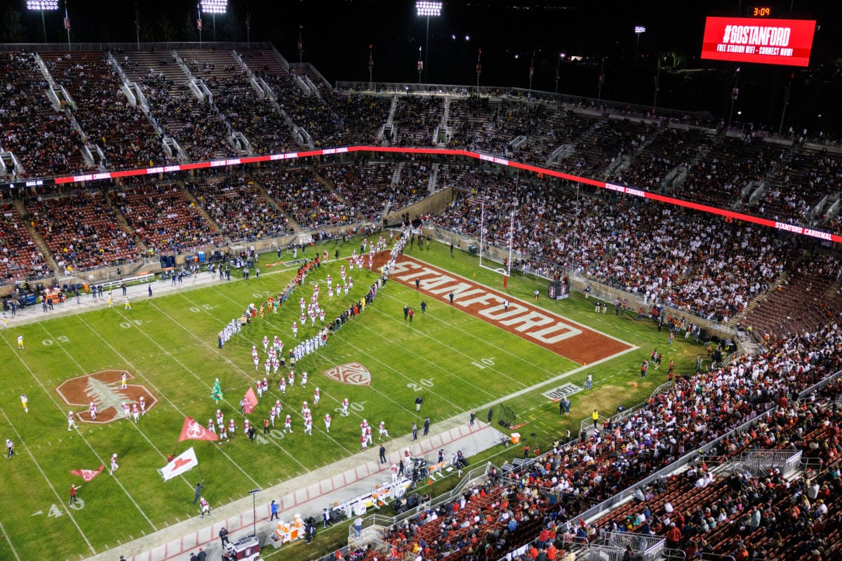 Wide shot from above of the Stanford football stadium, people in the bleachers, and Stanford football players emerging from the tunnel onto the field. 