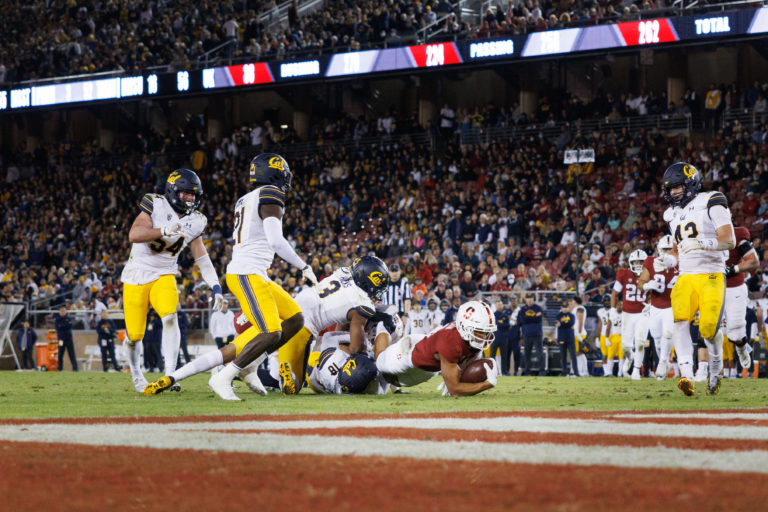 3 Cal football players piled on top of Stanford football player holding the football on field, just short of goal line