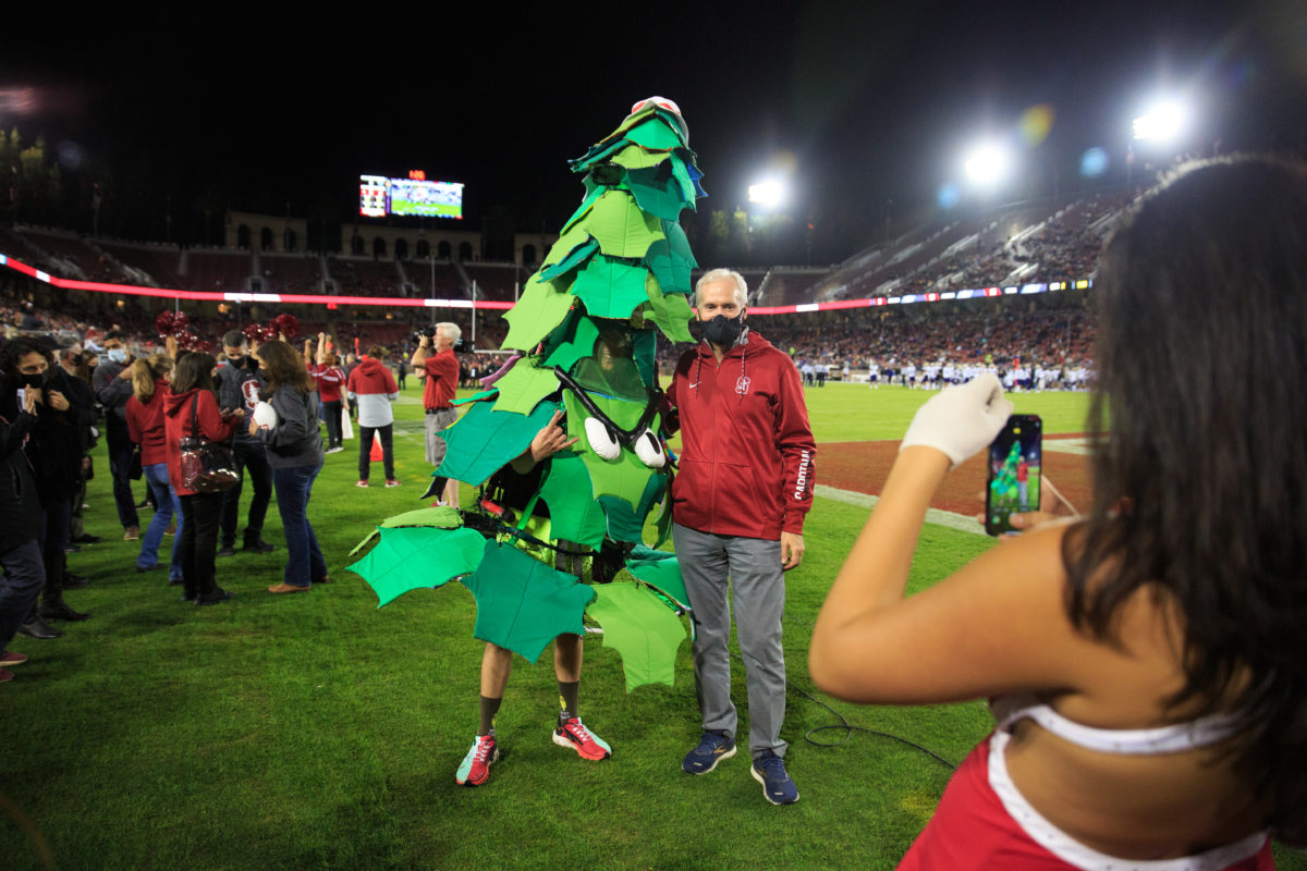 Stanford tree mascot next to Stanford President Marc Tessier-Lavigne on the football field. Their picture is being taken by a cheerleader.