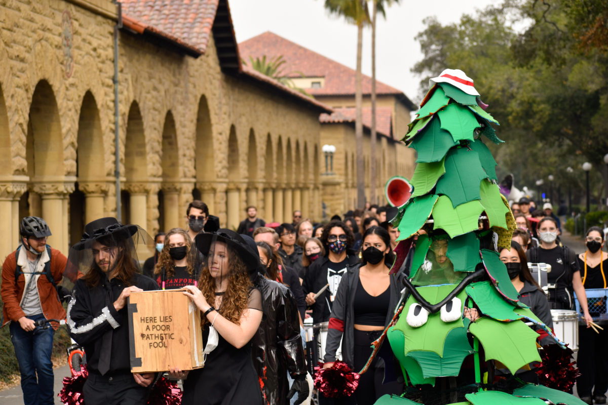 The Stanford Band, dollies, and tree dressed in black attire walk alongside Main Quad. Two band members hold a wooden casket, which reads "Here lies a poor pathetic fool".